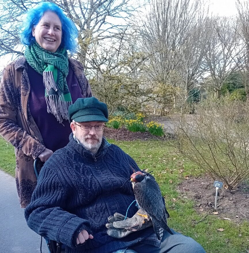 Dave is sat in his wheelchair with a bird on his lap. Kat is standing behind him, pushing his chair