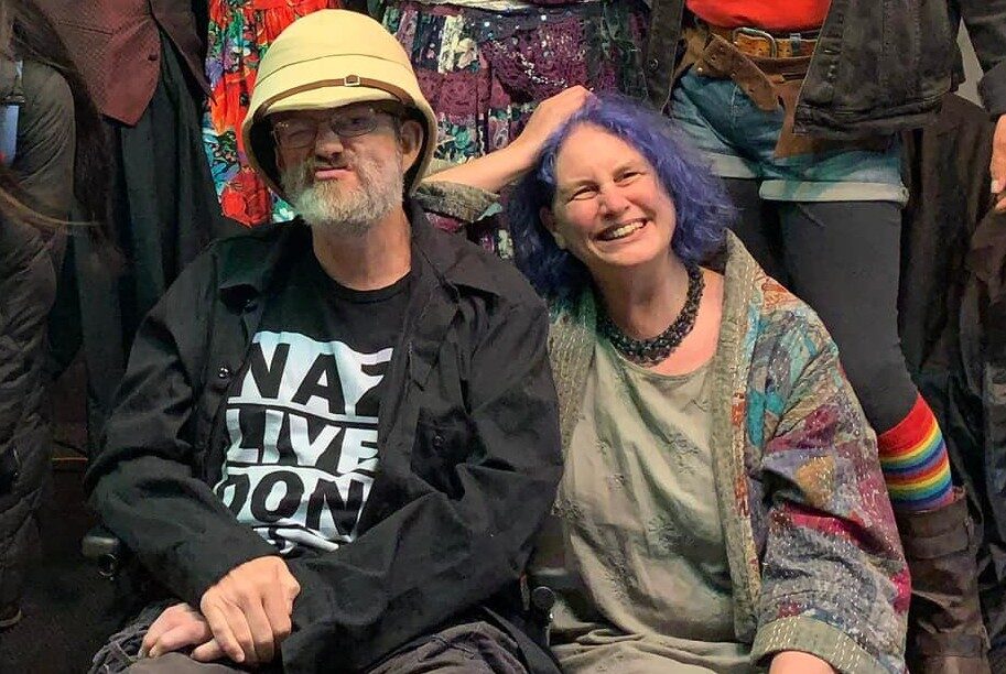 Man wearing a hat and black clothes is sat next to a woman with blue hair. She is smiling.